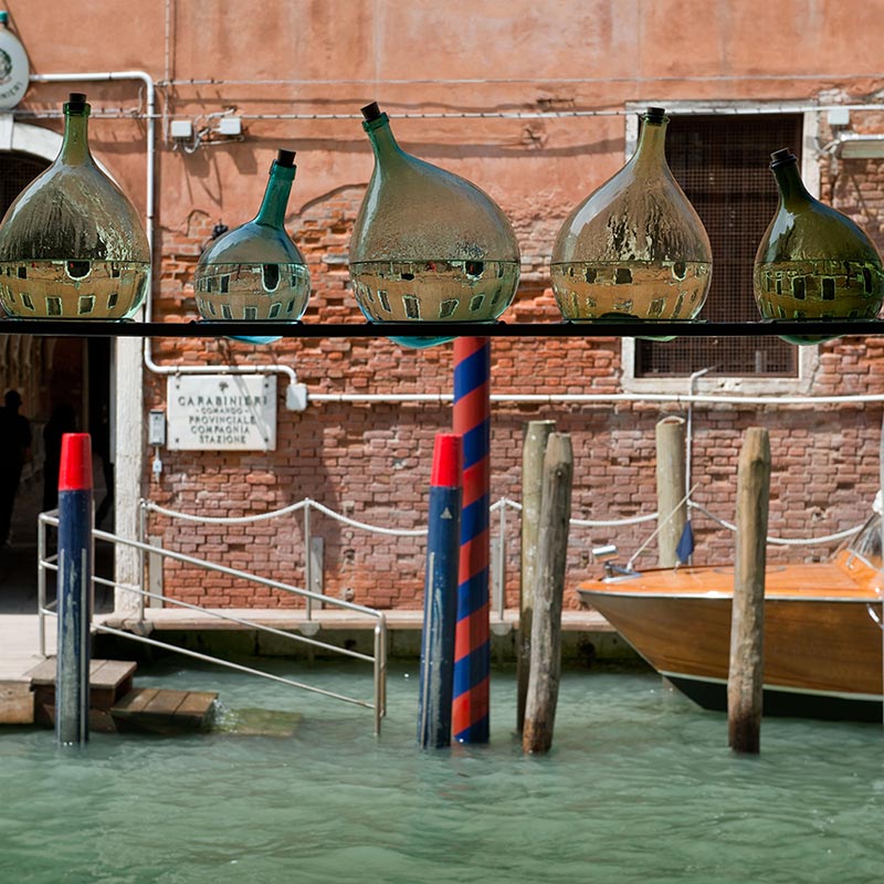 Six tear-shaped glass bottles in various sizes and containing water which creates a reflection of the red brick building on the opposite side of the canal, presented in a row across a steel shelf spanning the external doorway in front of the canal 
