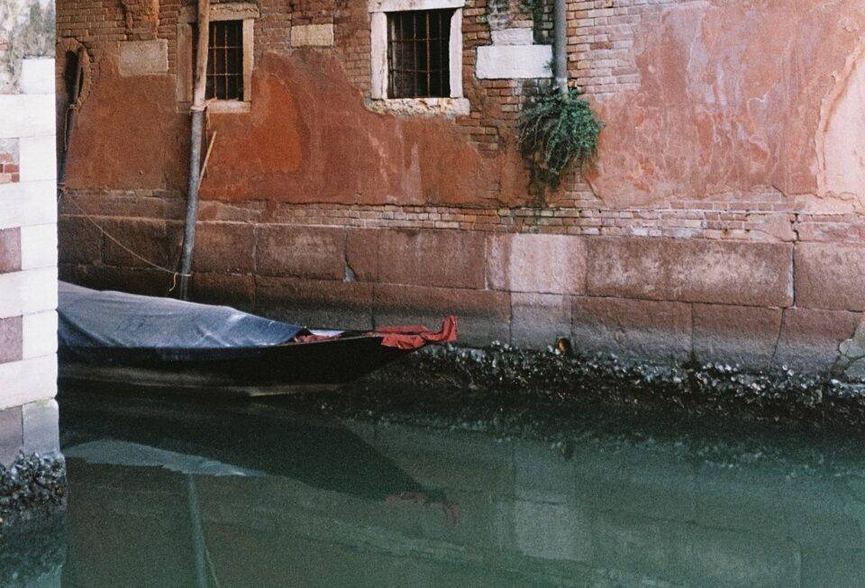 A black gondola sits stowed and covered at its mooring along a narrow canal. Image supplied.
